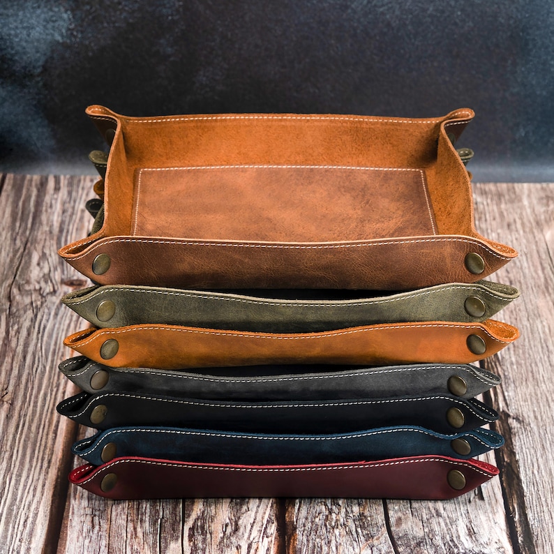 May include: A stack of rectangular leather trays in various colors, including brown, green, gray, and blue. Each tray has a contrasting white stitch and metal snap closures. The trays are stacked on a wooden surface.