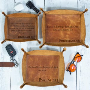 May include: Three brown leather catchall trays with rounded corners and scripture quotes. One tray has "JEREMIAH 29:11", another "PHILIPPIANS 4:13", and the third "PSALM 23:1". Accessories like sunglasses and a watch are visible.