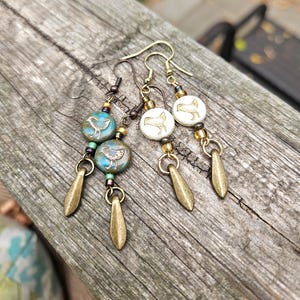 May include: Handmade earrings featuring bird-themed beads in turquoise and white, accented with gold-colored beads and teardrop charms. The earrings have a vintage aesthetic, with a rustic finish. The earrings are displayed on a wooden surface.