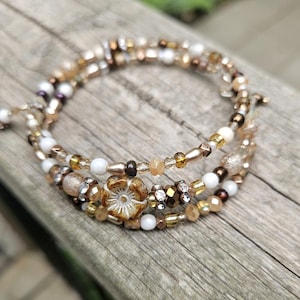 Peut inclure: Un bracelet à plusieurs brins de perles dans les tons or, marron et blanc. Le bracelet comporte un motif floral et diverses formes de perles, notamment rondes, à facettes et cylindriques. Le bracelet est enroulé sur lui-même.