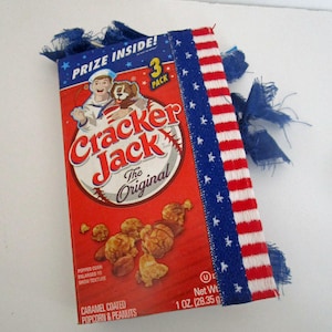 May include: A red Cracker Jack box with the original logo, featuring a sailor and dog illustration. The box is decorated with a blue ribbon with white stars and a red and white striped ribbon. The box contains 1 oz (28.35 g) of caramel-coated popcorn and peanuts.