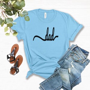 May include: A pale blue t-shirt with the word "dad" in black script. The shirt is styled with blue jeans and brown sandals with black straps. The shirt is knotted at the bottom.