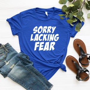 May include: A royal blue short-sleeved t-shirt featuring the white text SORRY LACKING FEAR printed in a bold, sans-serif font across the chest. The shirt is displayed alongside a pair of blue denim jeans and brown strappy sandals.