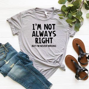 May include: A light gray t-shirt with the text "I'M NOT ALWAYS RIGHT BUT I'M NEVER WRONG" in black. The shirt is paired with blue jeans and brown sandals with black straps.