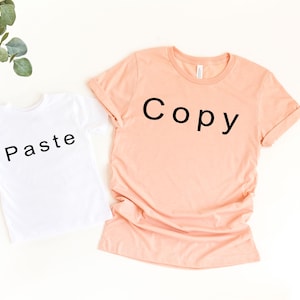 May include: Two t-shirts, one peach and one white, are displayed on a white surface. The peach shirt has the word "Copy" printed in black, while the white shirt has "Paste" printed in black. Green leaves are in the upper left corner.