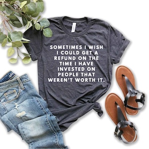 May include: A dark heather grey short-sleeve t-shirt with white text that reads: SOMETIMES I WISH I COULD GET A REFUND ON THE TIME I HAVE INVESTED ON PEOPLE THAT WEREN'T WORTH IT. The shirt is paired with blue denim jeans and brown sandals.