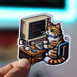 May include: A pixel art sticker of a tabby cat sitting at a computer desk with a cup of coffee. The cat is wearing a blue shirt and has a white collar. The computer screen shows code.