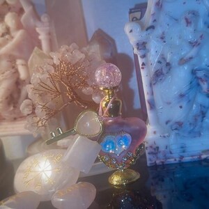 May include: A collection of decorative items, including a heart-shaped perfume bottle with a gold base and a crystal ball top. A key-shaped charm and a small crystal tree are also present. Two statues are in the background, one pink and one white.