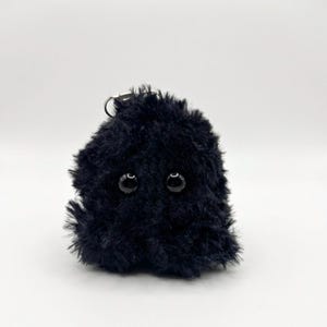 May include: A small, black, fuzzy creature with two large, round, black eyes. The plush toy has a keychain attached at the top. The background is plain white.