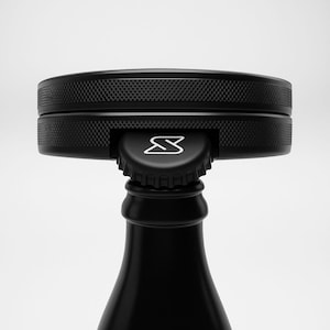 Puede incluir: Tapón de botella negro con una parte superior circular texturizada y un logotipo blanco. El tapón está diseñado para encajar en una botella, con una estética elegante y moderna. La botella es negra y tiene un acabado liso.