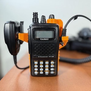 May include: A black Yaesu FT-60 FM transceiver with an orange mounting bracket and a black microphone. The radio has a digital display and a keypad with various function buttons. A black cable is connected to the radio.