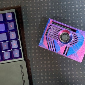 May include: A custom-built computer case with a vibrant pink, blue, and purple geometric design. The case features a central fan and ventilation slots. A backlit keyboard with purple keys is visible on the left.