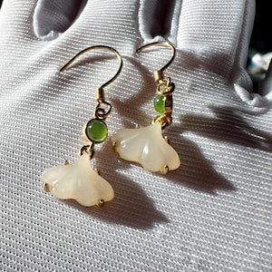 Puede incluir: Un par de pendientes dorados con un diseño en forma de pétalo de color claro. Cada pendiente tiene una pequeña piedra preciosa verde redonda y un cierre de gancho. Los pendientes se muestran sobre una superficie blanca.