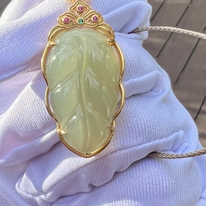 May include: A gold pendant with a leaf-shaped jade stone. The pendant has a gold frame and is adorned with small red and green gemstones. The jade is a pale green color and has visible veining. The pendant hangs from a thin, beige cord.