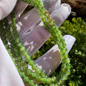 Op de afbeelding: Een close-up van een groene kralenketting met een gouden sluiting. De ketting is gemaakt van kleine, ronde, lichtgroene kralen. De sluiting is van goudkleurig metaal. De ketting wordt vastgehouden door een gehandschoende hand.