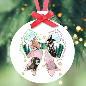 May include: White Christmas ornament with a pink ribbon and bow. The ornament features a bow design with a castle, witch hat, and forest scene. Text around the bow reads "Because I Knew You I Have Been Changed For Good."