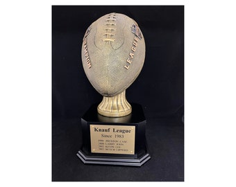 May include: A bronze-colored football trophy on a black base. The trophy is engraved with "Knauf League" and the years since 1983. The football has the words "Champion" and "League" on it.
