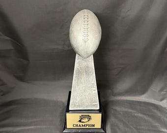 May include: A silver-colored football trophy with a black base and a gold-colored plate that reads "Fantasy Football Champion". The trophy is designed to resemble a football.