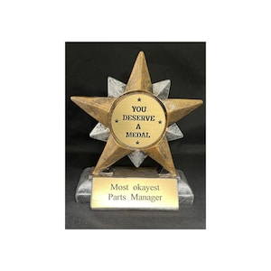 May include: Gold and silver star-shaped trophy with a round plaque that reads "You Deserve A Medal" and a base that reads "Most okayest Parts Manager".