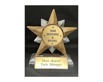 May include: Gold and silver star-shaped trophy with a round plaque that reads "You Deserve A Medal" and a base that reads "Most okayest Parts Manager".