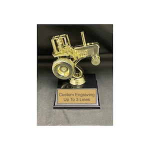 May include: A gold-colored tractor trophy on a black base. The trophy features a detailed tractor design with large wheels and a cab. The base has a gold plate with the text "Custom Engraving Up To 3 Lines".