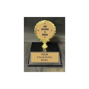 May include: A gold-colored trophy with a black base. The top of the trophy has a circular design with the words "YOU DESERVE A MEDAL" and stars. The base has a rectangular plate that reads "YOUR ENGRAVING HERE."
