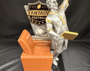May include: A silver-colored Fantasy Football trophy featuring a football player with his arm raised in victory. The trophy includes an orange chair, a football helmet, and a shield with the words "Fantasy Football".