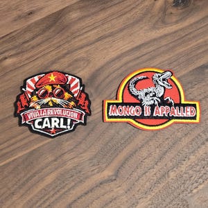 May include: Two embroidered patches. The left patch features a cat wearing a red beret and sunglasses with the text VIVA LA REVOLUCION CARL!. The right patch features a dinosaur skeleton inside a yellow and red circle with the text MONGO IS APPALLED.