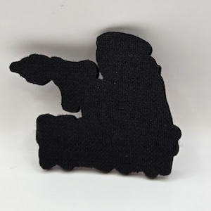 May include: A black, irregularly shaped patch with a textured surface. The patch has a silhouette design with a jagged bottom edge and a rounded top. The patch is set against a white background.