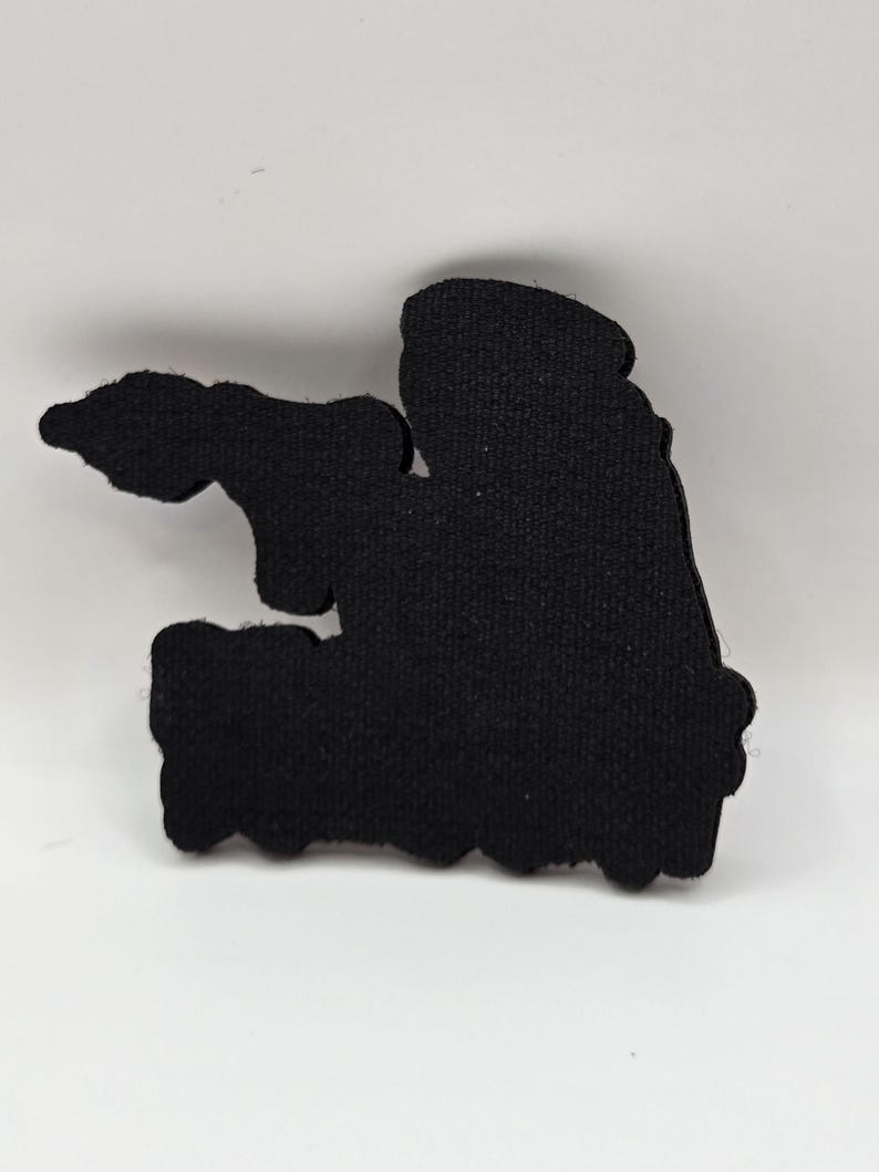 May include: A black, irregularly shaped patch with a textured surface. The patch has a silhouette design with a jagged bottom edge and a rounded top. The patch is set against a white background.