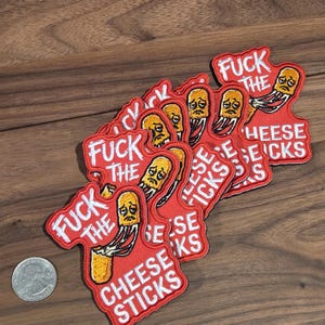 May include: A collection of embroidered patches featuring a red background with white text that reads FUCK THE CHEESE STICKS and an illustration of a sad yellow cheese stick with melting strands. A silver US quarter coin is placed for scale.