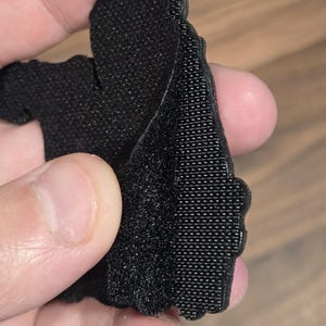 May include: Close-up of a black, textured fabric piece with a hook-and-loop fastener. The fabric is held up, revealing the texture and construction of the material. The edges are cut in a decorative pattern.