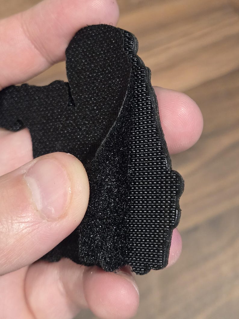 May include: Close-up of a black, textured fabric piece with a hook-and-loop fastener. The fabric is held up, revealing the texture and construction of the material. The edges are cut in a decorative pattern.