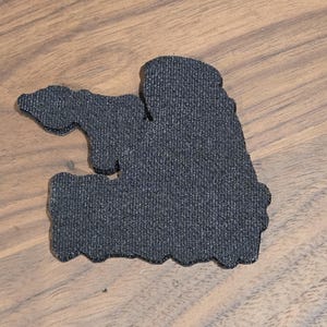 May include: A dark gray, textured patch with an irregular shape. The patch has a rough, fabric-like appearance and is set against a wooden surface.