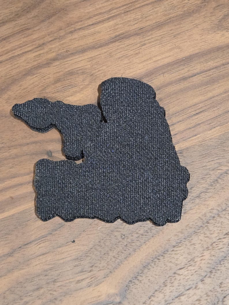 May include: A dark gray, textured patch with an irregular shape. The patch has a rough, fabric-like appearance and is set against a wooden surface.