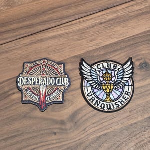 May include: Two embroidered patches on a wooden surface. One patch reads "DESPERADO CLUB SO FUN IT HURTS" with a red and white design. The other patch says "CLUB VANQUISHER" with a winged design and a chalice.