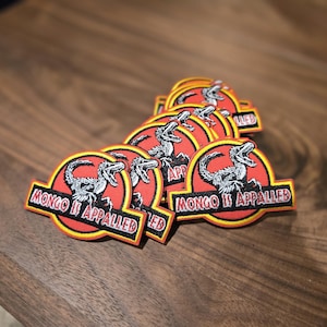 Puede incluir: Un montón de parches bordados con un diseño circular rojo y amarillo. Los parches presentan un gráfico de dinosaurio y el texto "MONGO IS APPALLED". Los parches están sobre una superficie de madera oscura.