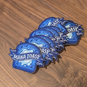 Puede incluir: Un conjunto de parches bordados azules con forma de rebanada de pan. Cada parche tiene las palabras "MANA TOAST" en una pancarta. El pan tiene un diseño azul y blanco, y los parches están colocados sobre una superficie de madera.