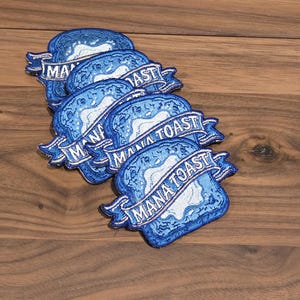 Puede incluir: Una colección de cuatro parches bordados con forma de rebanadas de pan tostado, con corteza azul y centro blanco. Cada parche incluye una cinta azul en la parte frontal con el texto MANA TOAST bordado en letras mayúsculas blancas.