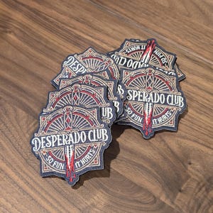Puede incluir: Un montón de parches bordados con la frase "DESPERADO CLUB". Los parches presentan un diseño rojo y blanco con el texto "SO FUN IT HURTS" y un gráfico de brújula. Los parches están sobre una superficie de madera.