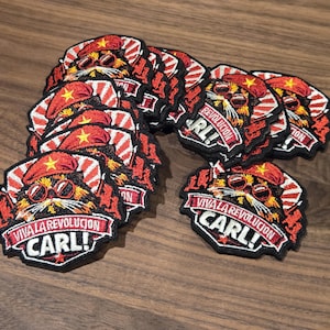 Peut inclure: Plusieurs écussons brodés avec un chat portant des lunettes de soleil et un béret rouge. Les écussons comportent le texte "VIVA LA REVOLUCION CARL!" et "REVOLUCION" en rouge et blanc.