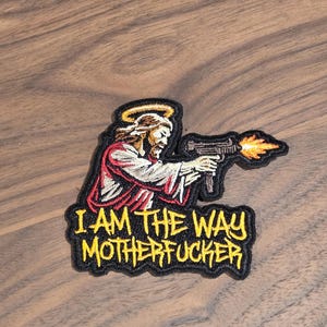 May include: Embroidered patch featuring Jesus with a halo, holding a gun with flames. The text "I AM THE WAY MOTHERFUCKER" is in yellow on a black background. The patch is on a wooden surface.