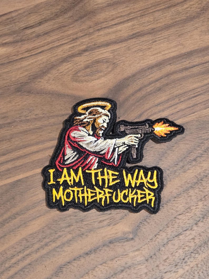 May include: Embroidered patch featuring Jesus with a halo, holding a gun with flames. The text "I AM THE WAY MOTHERFUCKER" is in yellow on a black background. The patch is on a wooden surface.
