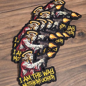 Op de afbeelding: Geborduurde patches met een afbeelding van Jezus Christus die een machinegeweer vasthoudt, met een halo en de tekst "I AM THE WAY MOTHERFUCKER" in geel. De patches hebben een zwarte rand en zijn gestapeld op een houten oppervlak.