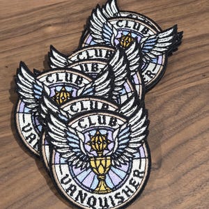 May include: A stack of embroidered patches with a circular design. Each patch features a winged emblem with the words "CLUB VANQUISHER" in a circular pattern. The patches are in shades of black, white, gold, purple, and blue.