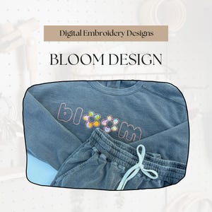 May include: A blue-grey sweatshirt and matching shorts set. The sweatshirt features the embroidered text "bloom" with flower designs. The text "Digital Embroidery Designs" and "BLOOM DESIGN" are displayed above the clothing.