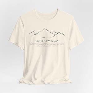 May include: Cream-colored t-shirt featuring a minimalist mountain range design in black ink. The text "MATTHEW 17:20" is above a quote about faith. The shirt is made of soft material and is suitable for casual wear.