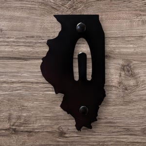 May include: A black metal wall hanging in the shape of the state of Illinois, with a cutout and two black circular fasteners. The wall decor is mounted on a wooden surface.