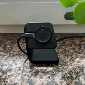 May include: A black Amazfit smartwatch charging dock. The dock has a circular charging pad with the Amazfit logo and a rectangular base. A black cable is connected to the dock.