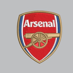 May include: A shield-shaped embroidered patch featuring the Arsenal football club logo. The design includes a red background with white text reading Arsenal, a gold cannon graphic, and a gold border with blue and white inner trim details.